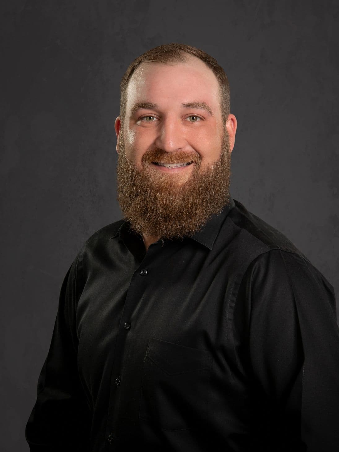Smiling man with a full beard wearing a black shirt against a dark background.