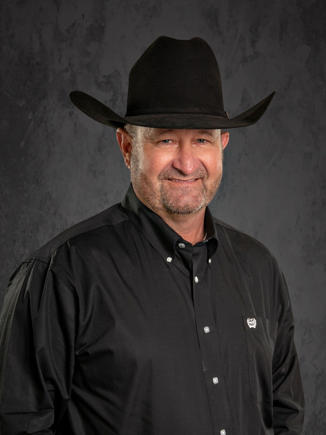 A man in a black cowboy hat and shirt smiling at the camera.