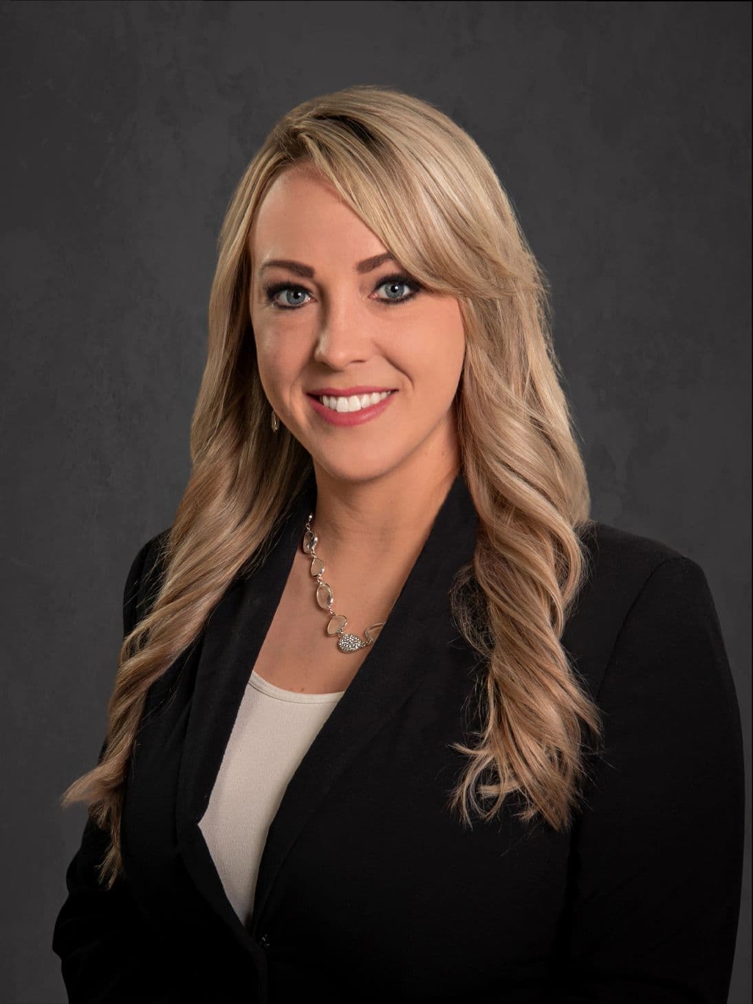 Professional woman with blonde hair in a black blazer smiling against a dark background.